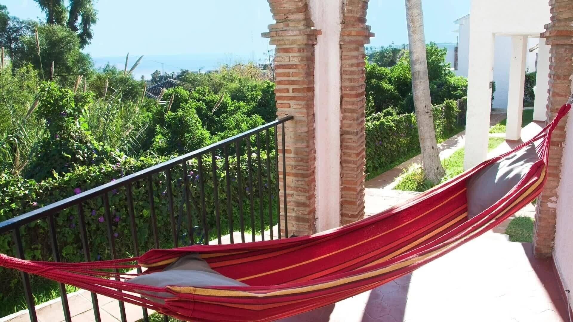 Un hamac sur un balcon, avec vue sur la verdure et un bâtiment en arrière-plan.