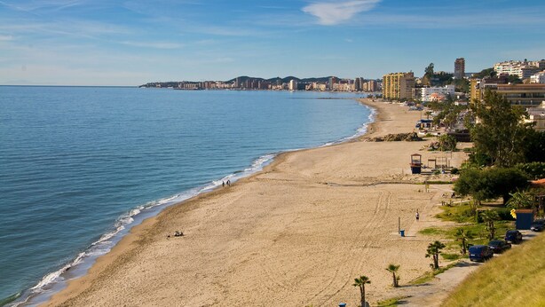 A coastal cityscape with a wide sandy beach, a clear blue sky, and buildings along the shore.