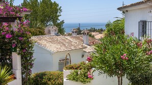 A coastal building with terracotta tiled roofs, white walls, and balconies with flowering plants.