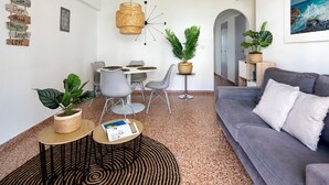 A modern living room with a grey sofa, a round coffee table, and a dining area with a small table and chairs. There are potted plants and a wall clock. - Holiday Beach - Denia, Costa Blanca (Denia)