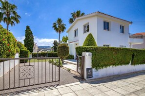 A white villa with a gated entrance, a well-maintained hedge, and a view of the surrounding landscape.