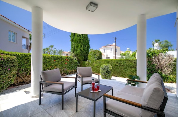Un espace extérieur aménagé, abrité et doté de deux chaises et d’une table, entouré de verdure et offrant une vue sur un bâtiment blanc.