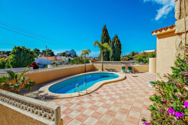 A swimming pool with a tiled deck, surrounded by greenery and a clear blue sky.