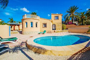 A swimming pool with lounge chairs, a stone building with arched windows, and palm trees.