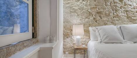 Une chambre avec un mur en pierre, un lit avec une literie blanche, une table de chevet avec une lampe et une fenêtre donnant sur des arbres.