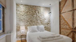 Un dormitorio con pared de piedra, una cama con sábanas blancas, una mesita de noche con lámpara y una ventana con vistas.