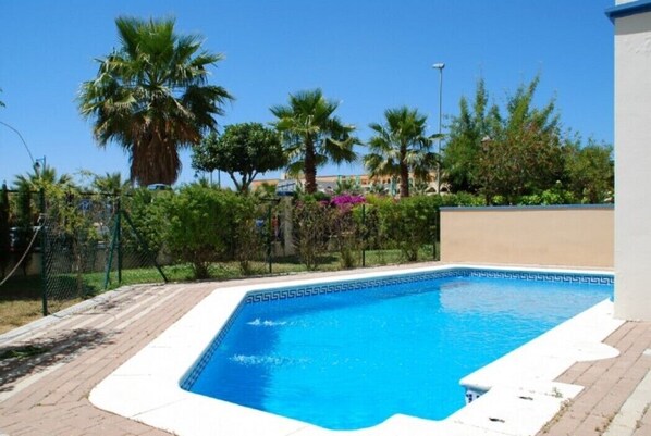 A rectangular swimming pool with a white edge, surrounded by a brick walkway and palm trees.