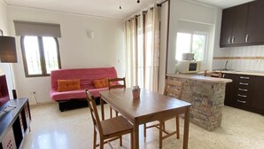 Ein kompakter Wohnbereich mit Küchenzeile, einer Essecke mit Holztisch und -stühlen, einem pinkfarbenen Sofa mit orangefarbenen Kissen sowie einem Deckenventilator.