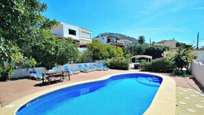 Una piscina con lettini e ombrellone, circondata dal verde e da edifici.