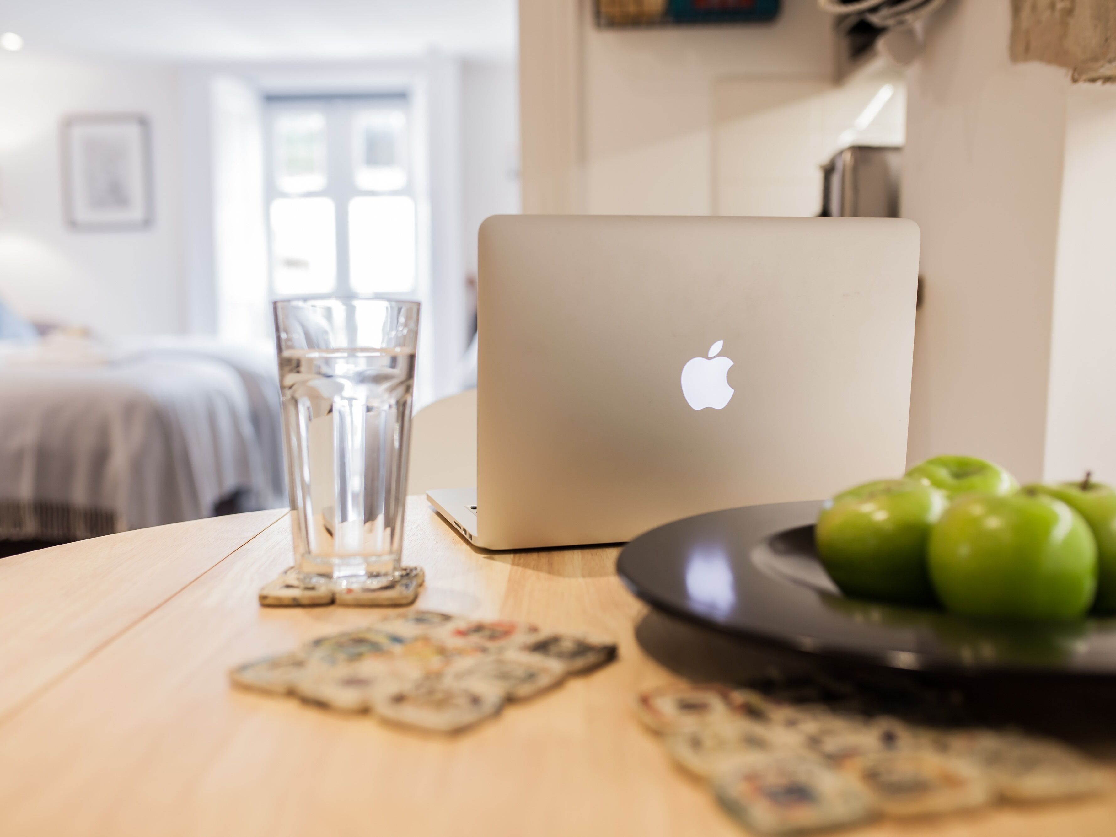 A laptop on a table with a glass of water, a plate of green apples, and some coasters.