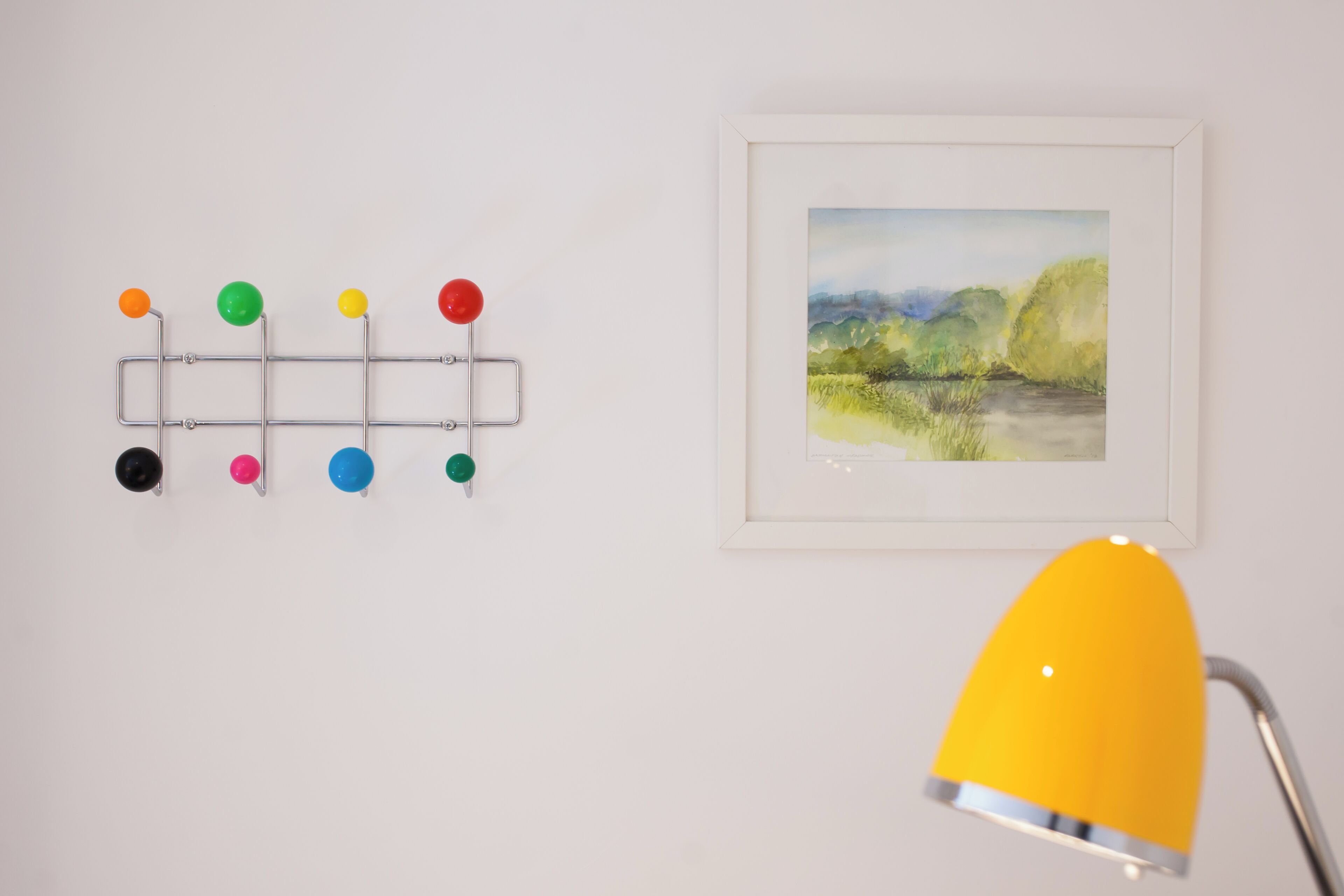 Un crochet mural orné d’épingles colorées, un tableau représentant un paysage encadré et une lampe de bureau jaune.