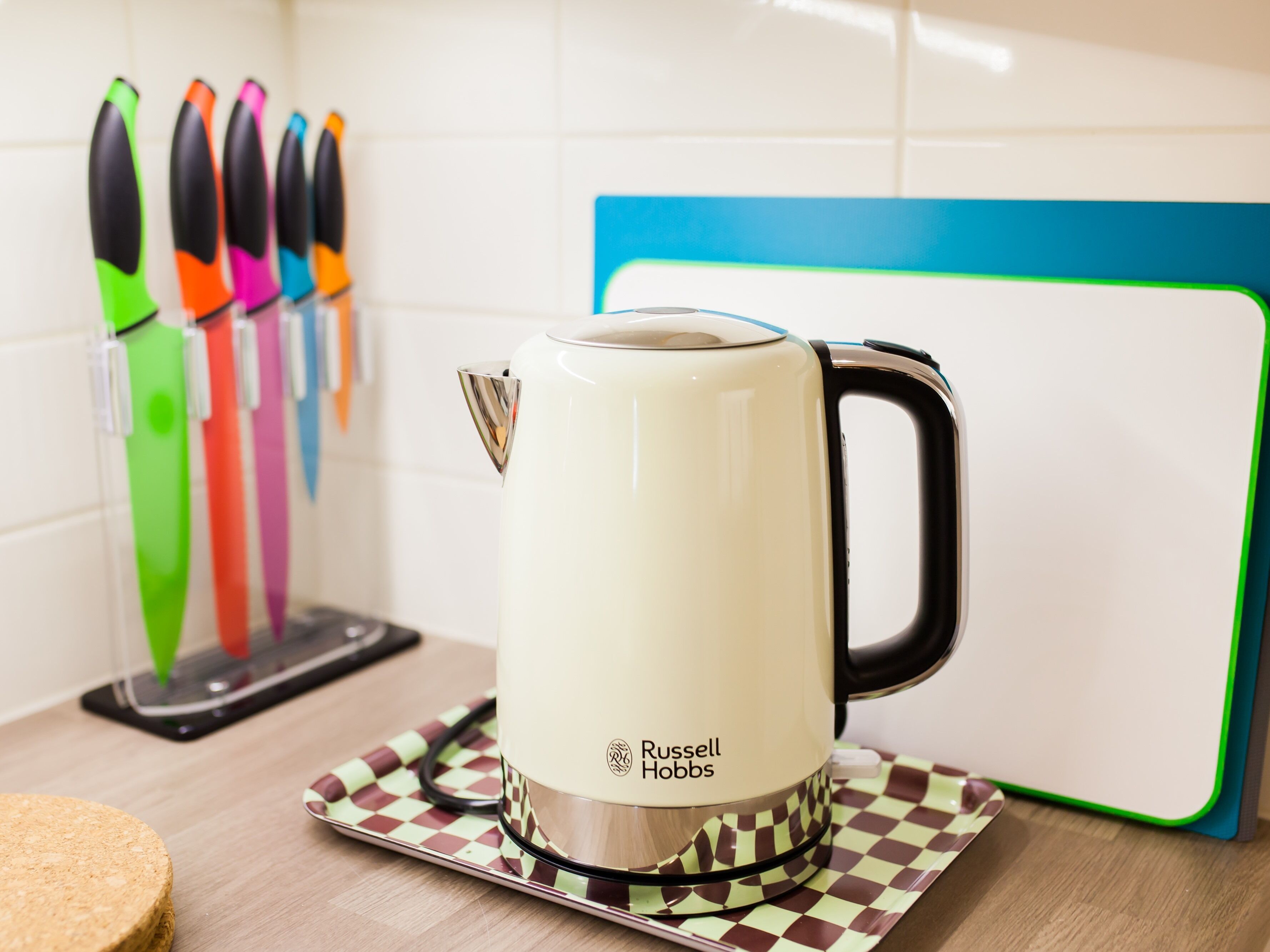 Una cocina con una tetera Russell Hobbs blanca sobre un posavasos de cuadros, cuchillos de colores en un portalápices y una servilleta de mesa azul y verde.