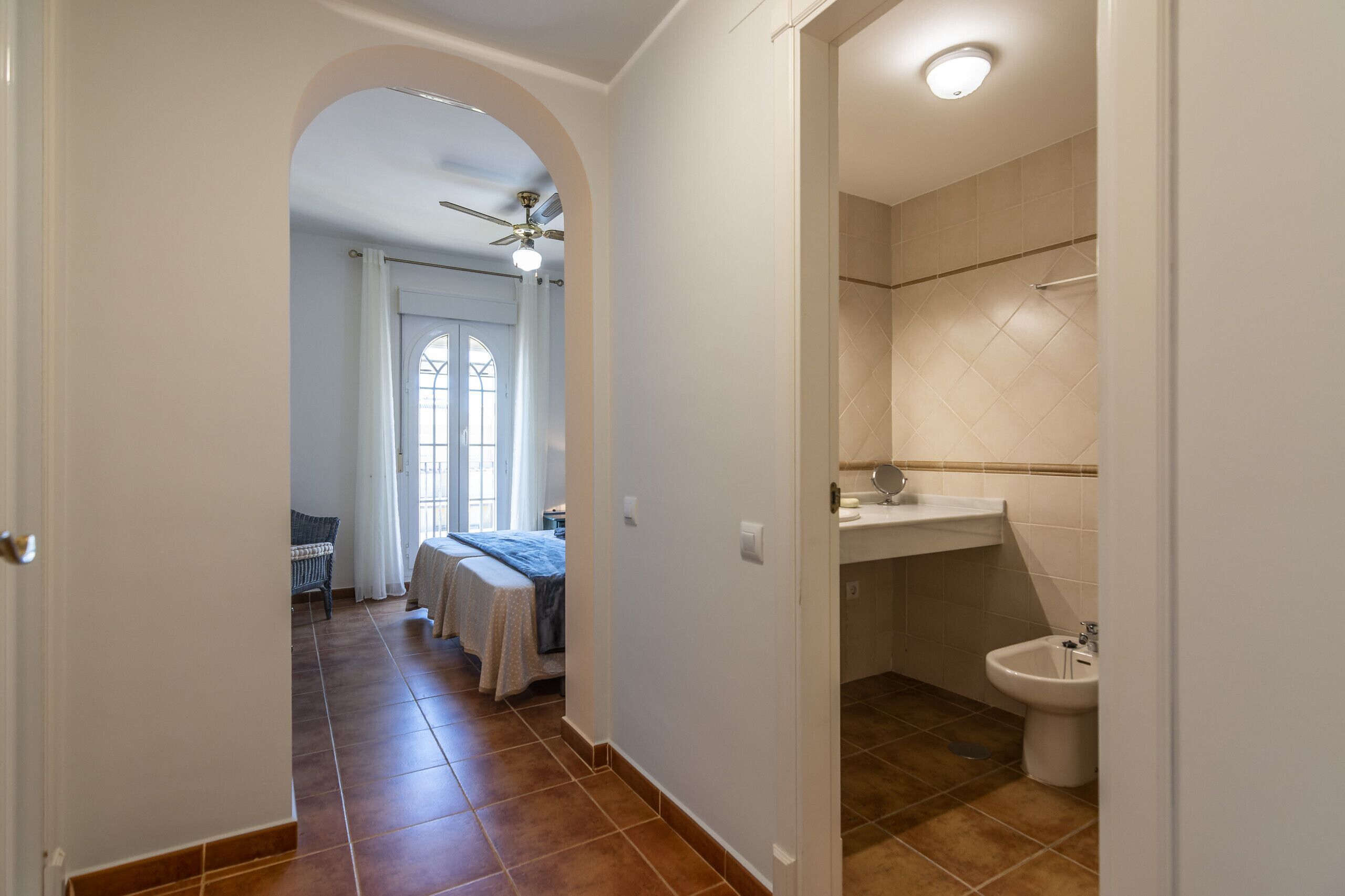 Un couloir mène à une chambre dotée d’un ventilateur de plafond, d’une fenêtre avec des rideaux et d’un lit recouvert d’une couverture bleue. La salle de bain comprend des toilettes, un lavabo et un espace douche.