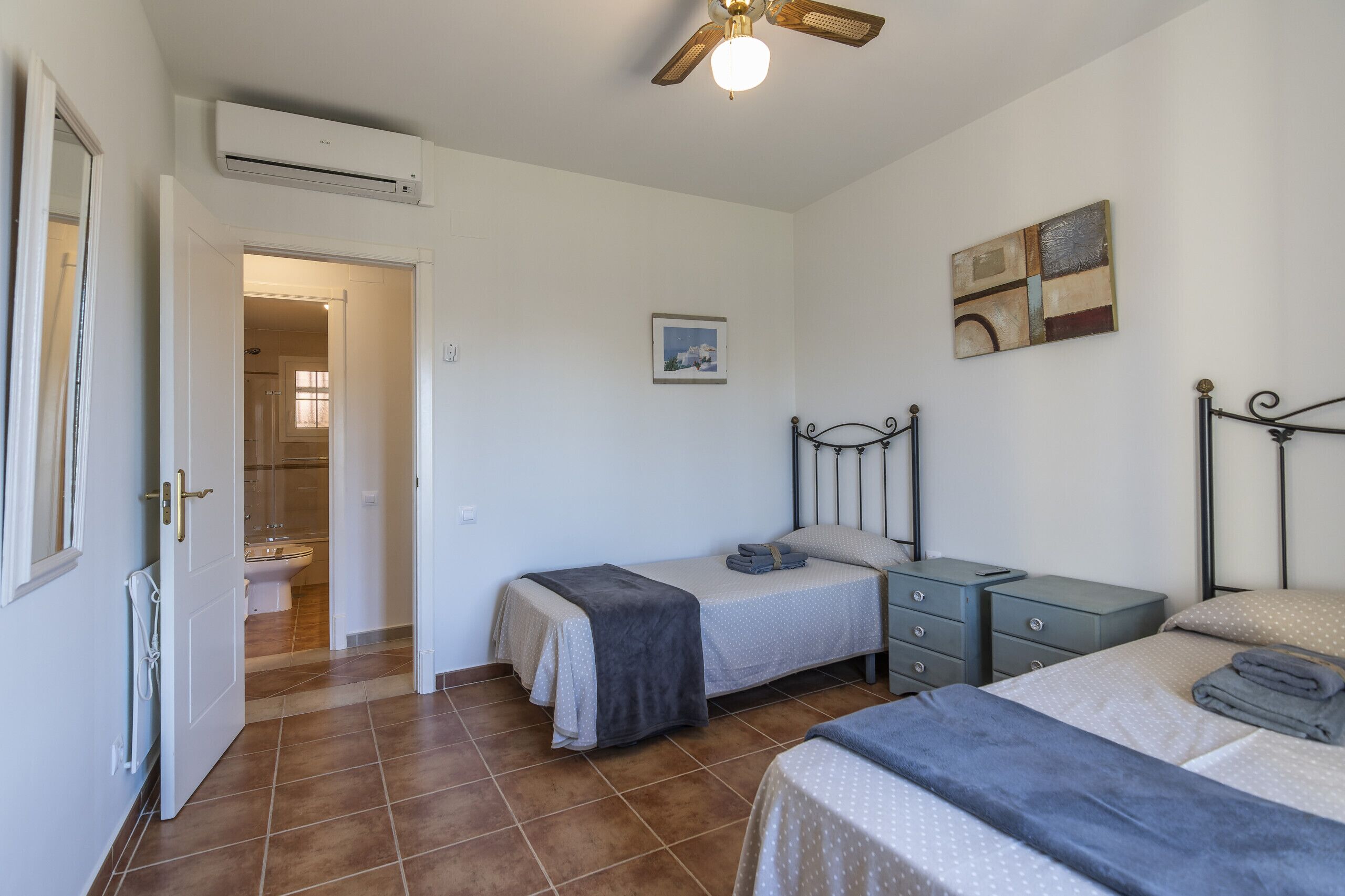 Quarto com duas camas, uma mesa de cabeceira, ar condicionado, ventoinha no teto e uma porta para o banheiro.