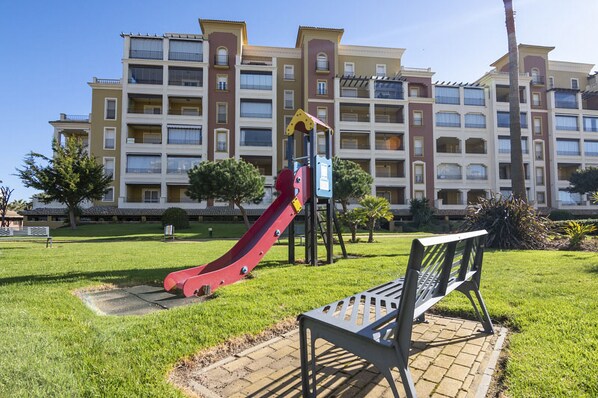 Un parque infantil con un tobogán rojo y un banco frente a un edificio de apartamentos de varios pisos.