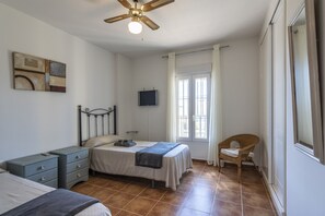 Un dormitorio con cama, mesita de noche, silla, ventilador de techo, televisor y ventana con cortinas.