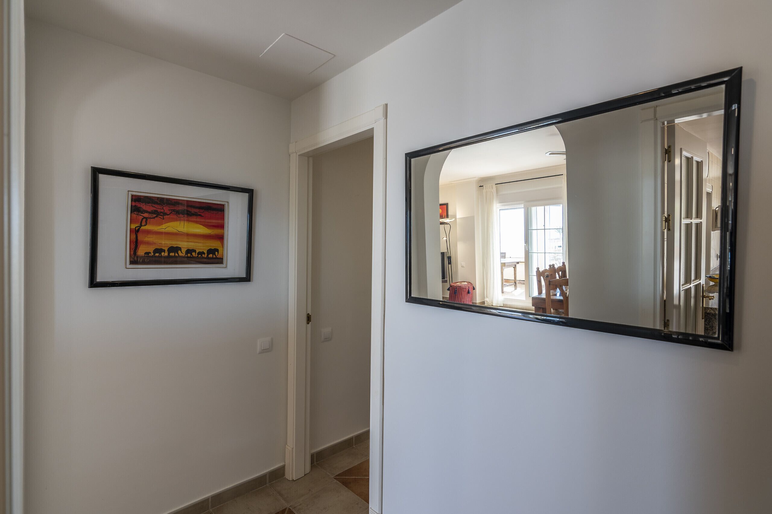 A hallway with a framed painting, a mirror, and a door leading to another room.