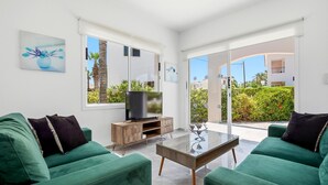 A modern living room with green sofas, a glass coffee table, a TV on a wooden cabinet, and large windows offering a view of greenery and buildings. - Villa Shiva by Ezoria Villas (Coral Bay)