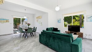 A spacious living area with a green sofa, dining table, and chairs, a flat-screen TV, and artwork on the walls. - Villa Shiva by Ezoria Villas (Coral Bay)