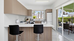A modern kitchen with a central island, two bar stools, a view through the window, and a balcony with outdoor furniture. - Villa Shiva by Ezoria Villas (Coral Bay)