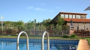 Una piscina con escaleras de metal en primer plano, una valla de madera y una casa con techo de tejas al fondo.