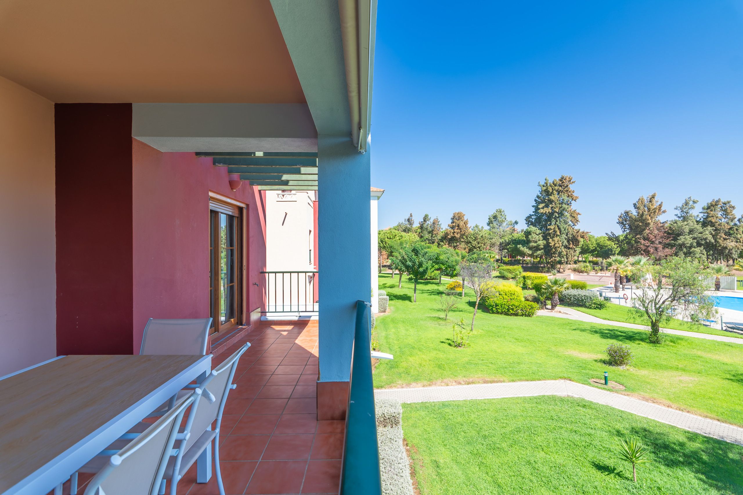 Un balcón con mesa y sillas, vista a un jardín cuidado y un cielo azul despejado.