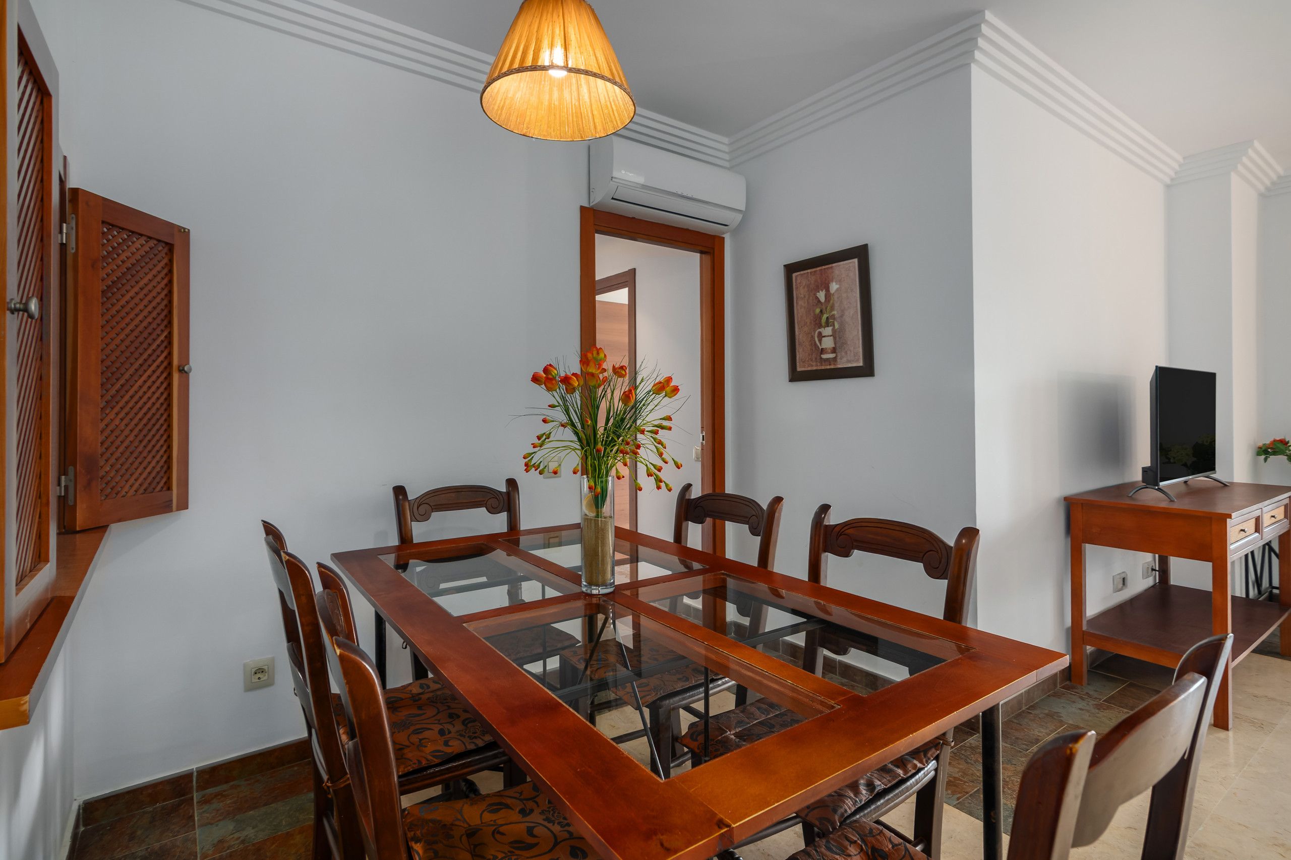 Un comedor con una mesa de madera, sillas, un jarrón con flores, una lámpara colgante y un televisor sobre un mueble de madera.