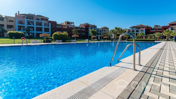Una piscina con acqua cristallina e blu, circondata da un solarium piastrellato e una scaletta in metallo.