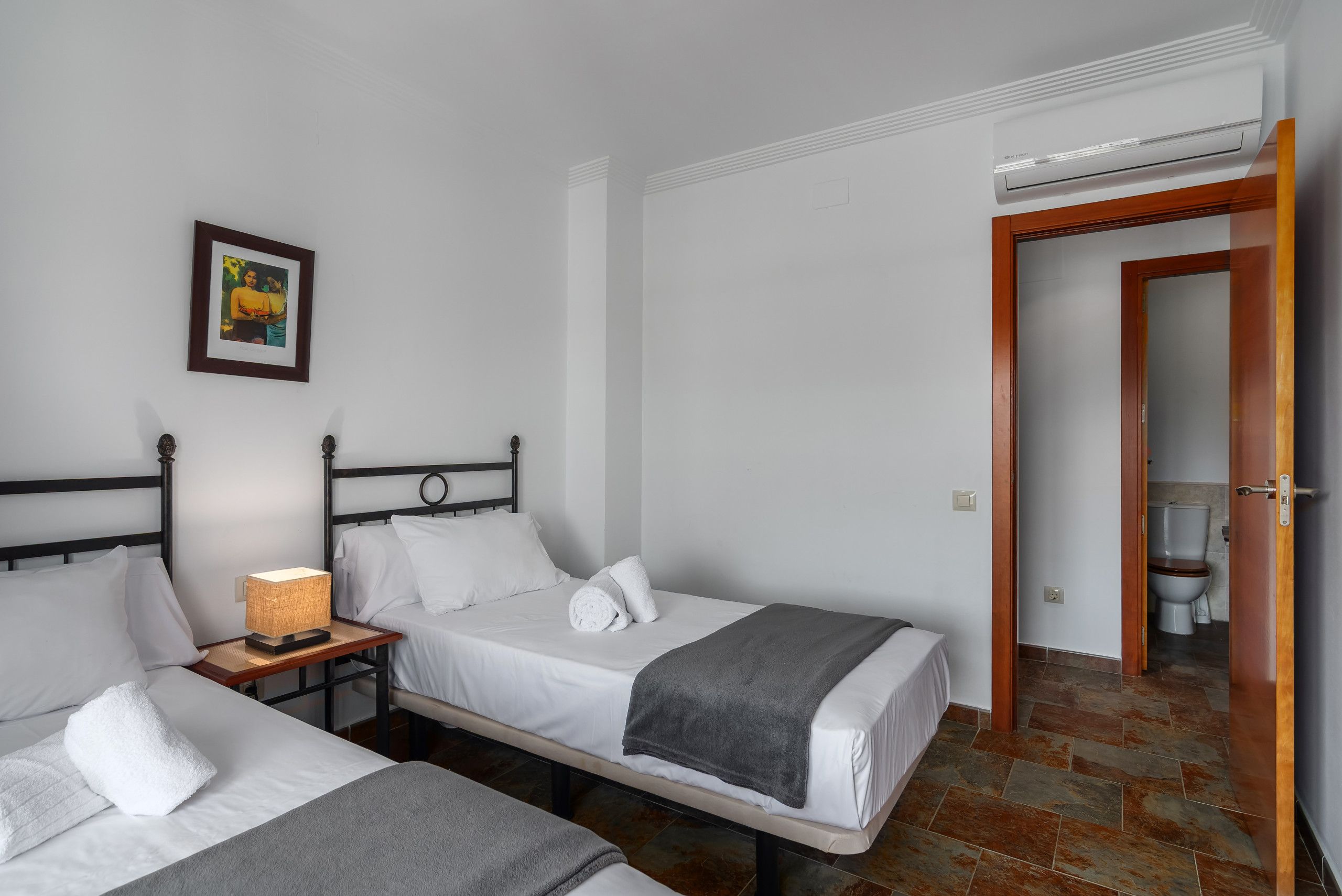 Habitació d'hotel amb dos llits individuals, una taula de nit amb una làmpada, un quadre amb marc a la paret i un bany visible a través d'una porta oberta.