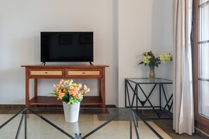 A room with a wooden console table, a flat-screen TV mounted above it, a glass coffee table with a vase of flowers, and a small side table with a vase of flowers. - El Rincon del Golf III PLUS - Isla Canela Golf GRHIII88P88 (Isla Canela)