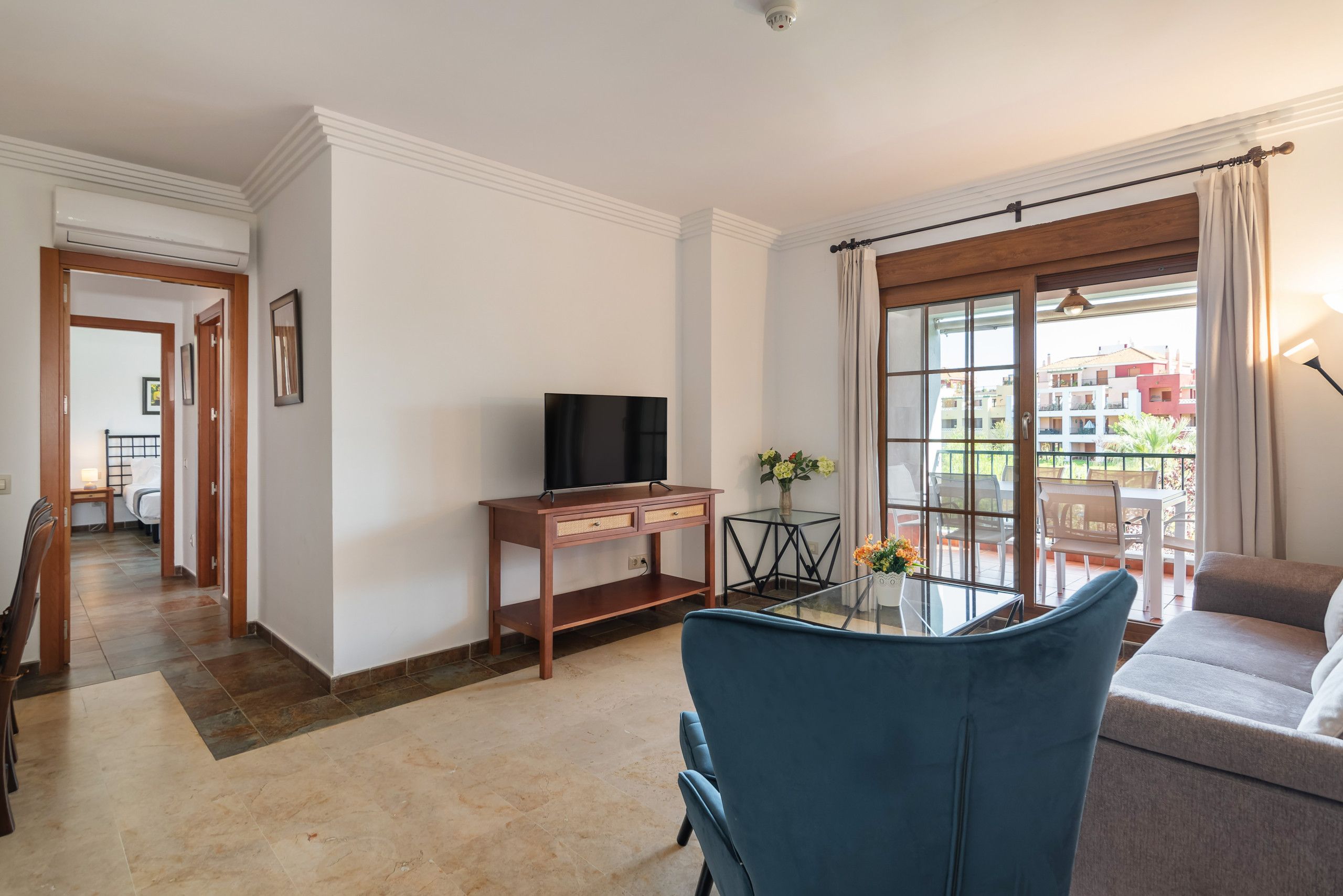 Ein Wohnzimmer mit Fernseher, einem Glastisch und einer Couch. Es gibt einen Balkon mit Blick auf Gebäude.