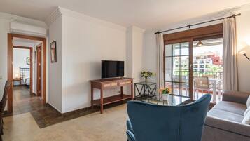 Ein Wohnzimmer mit Fernseher, einem Glastisch und einer Couch. Es gibt einen Balkon mit Blick auf Gebäude.