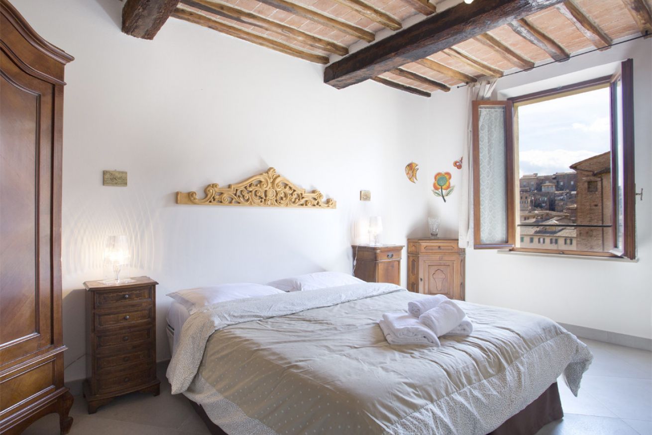 A bedroom with a bed, wooden dresser, nightstands, a decorative headboard, and a window with a city view.