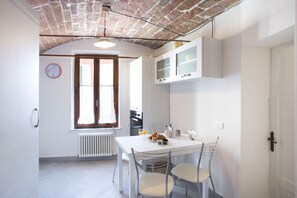 A compact kitchen with white cabinets, a dining table with chairs, a clock on the wall, and a window with curtains.