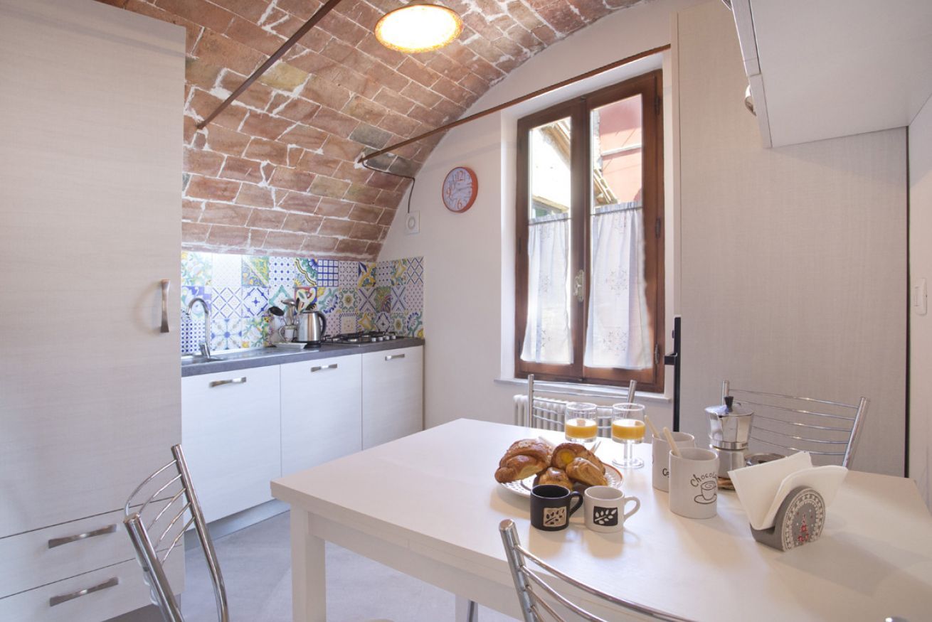 Una cocina con armarios blancos, salpicadero de azulejos y una ventana con vistas. Hay una mesa con cruasanes, tazas de café y una balanza.