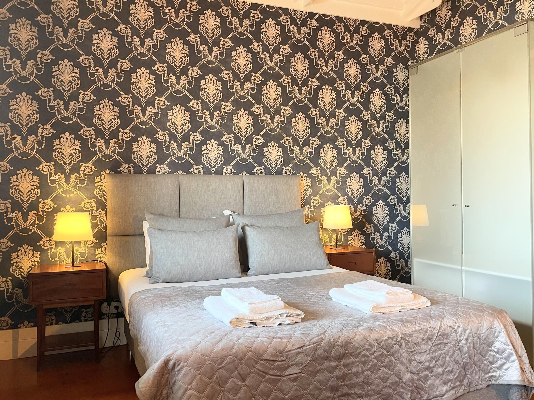 A neatly made bed with a quilted cover, two bedside lamps, and a folded towel on the bed.