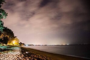 Een nachtelijk strandzicht met een rustige zee, een weg parallel aan de kust, en een boom met verlichte lampjes.