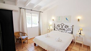 Ein Schlafzimmer mit einem Bett, zwei Nachttischen mit Lampen, einem Sessel, einem Fenster mit Vorhängen und Wandbildern.
