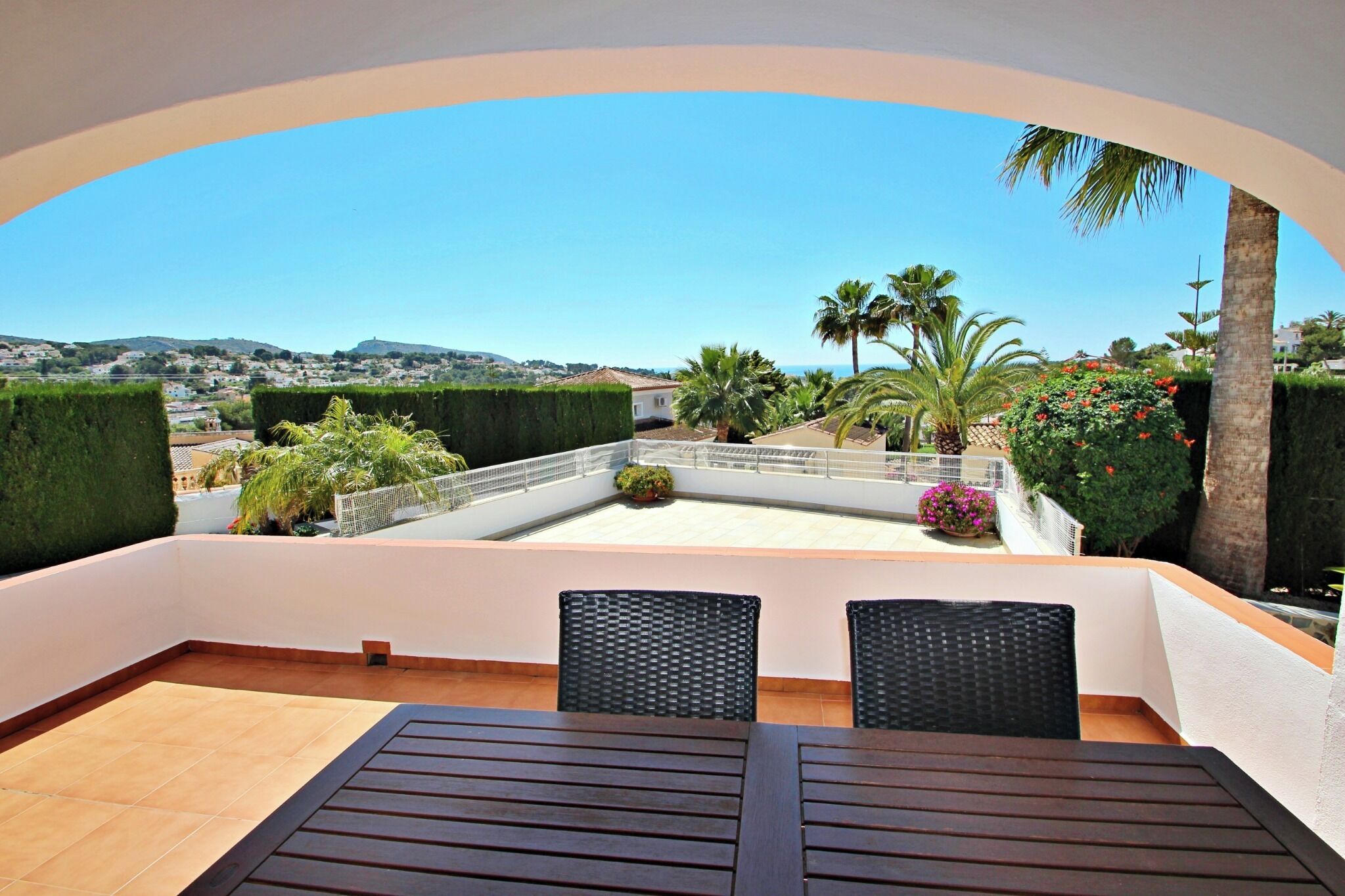 A rooftop terrace with a clear view of the cityscape, featuring a table and chairs, and surrounded by greenery and palm trees.