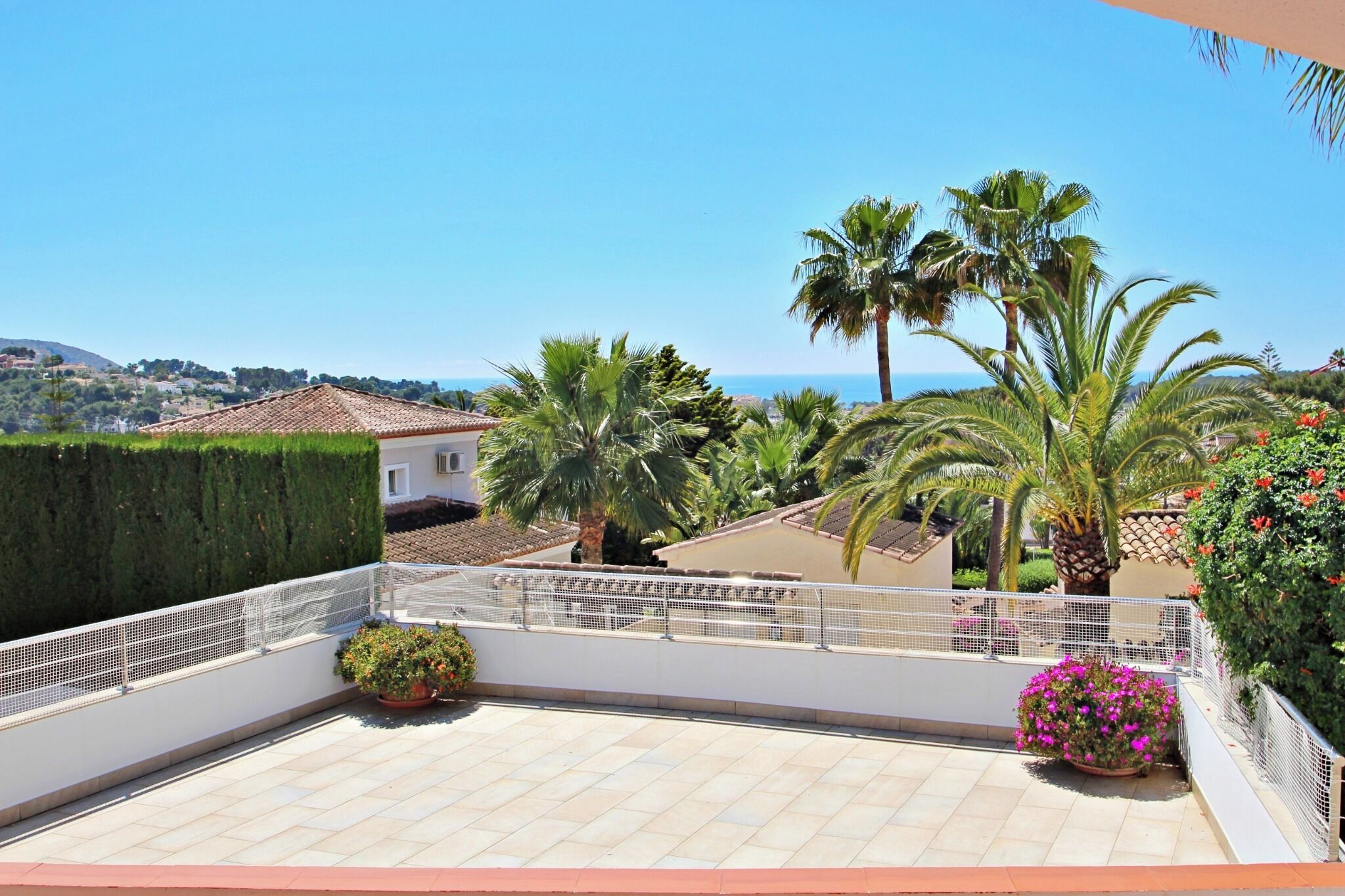 A rooftop terrace with a view of the sea, palm trees, and a white building.