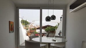 Un comedor moderno con una mesa redonda y sillas, un amplio ventanal corredero que da a un balcón con plantas y vistas a edificios.