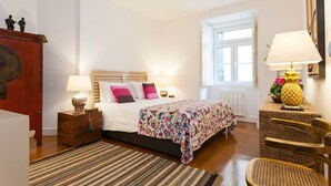 Un dormitorio con cama, lámparas de noche, cabecera de madera, ventana, mesita de mimbre y una silla.