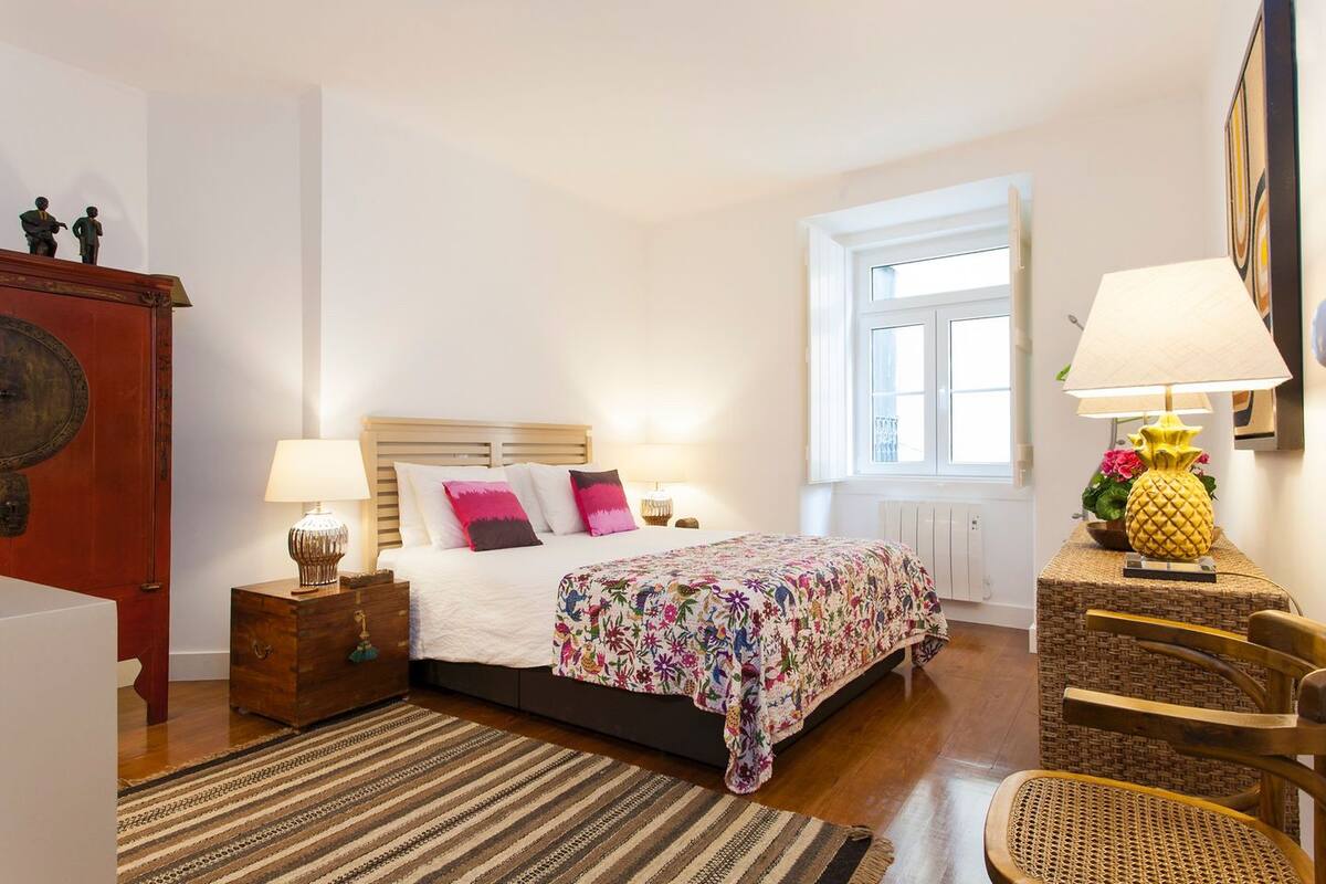 Une chambre à coucher comprenant un lit, des lampes de chevet, une tête de lit en bois, une fenêtre, une table d’appoint en osier et une chaise.