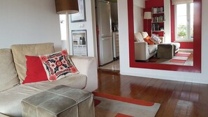Un salón con un sofá, un cojín estampado en rojo y negro, una lámpara y una estantería.