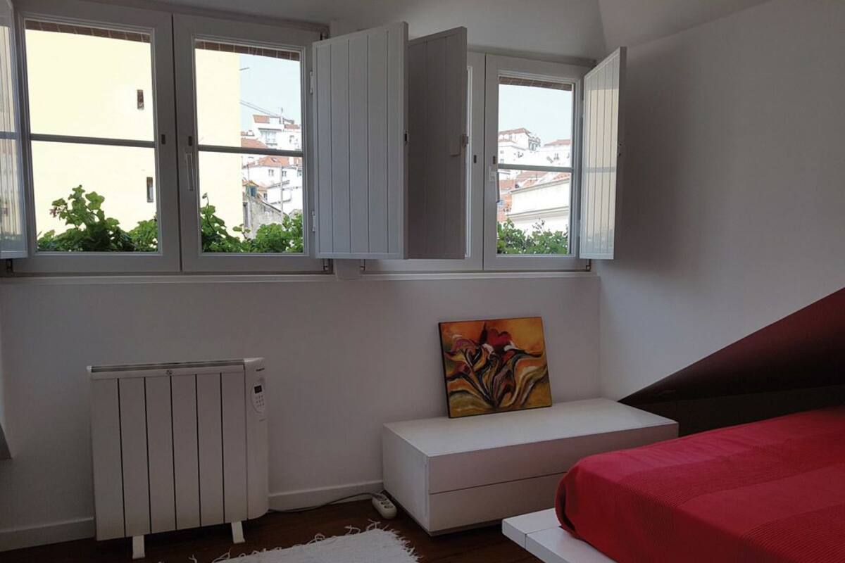 Une chambre à coucher avec un lit, un radiateur blanc, un banc blanc et une fenêtre dont les volets sont ouverts.