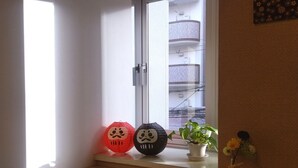 A window with a view of a building, a potted plant, and a small table with a teapot and a bottle. - Hostel Furoya (Osaka)