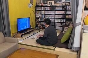 A person sitting on a tatami mat in a room with a television, a bookshelf, and a futon bed. - Hostel Furoya (Osaka)