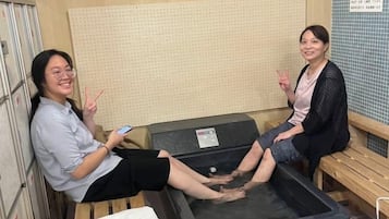 Two individuals seated in a room with a large, dark-colored, rectangular object in the center, possibly a sauna or hot tub, and a wooden bench to the right.