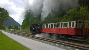 A steam train with red and white carriages traveling on tracks alongside a paved path, surrounded by greenery and a mountain in the background.