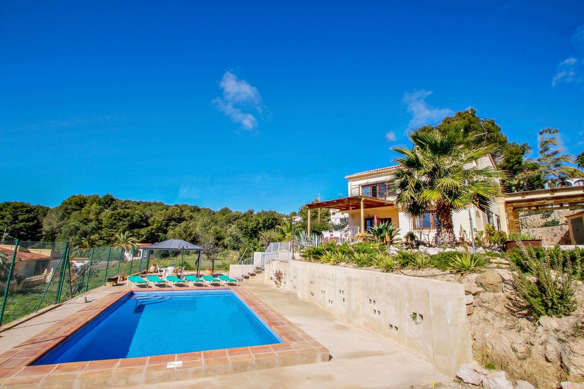 A swimming pool with lounge chairs, a stone wall, and a house with a patio.